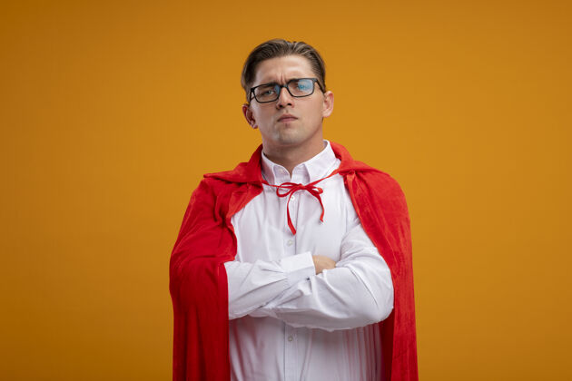胸部身穿红色斗篷 戴着眼镜 双手交叉放在胸前的超级英雄商人站在橙色的墙上脸站看