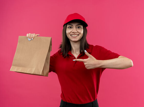举行身穿红色马球衫 头戴帽子的年轻送货女孩拿着纸包 食指指着纸包 微笑着站在粉红色的墙上 脸上洋溢着幸福目录马球脸