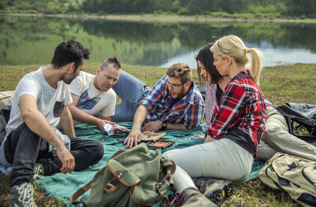 友谊一群年轻的朋友在阳光明媚的日子里坐在湖边的草地上背包湖泊团队