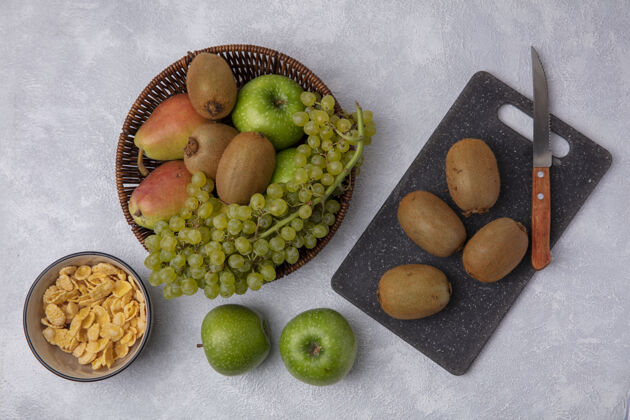 绿色顶视图猕猴桃用刀放在砧板上 绿色的苹果 葡萄和梨放在篮子里 玉米片放在碗里 背景是白色的碗视图食物