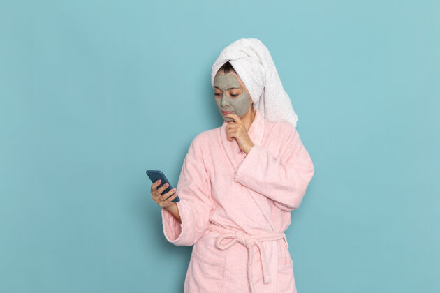 视图正面图年轻女性穿着粉色浴袍淋浴后用手机在浅蓝色墙面上美容水自理淋浴干净实验室外套淋浴浴袍