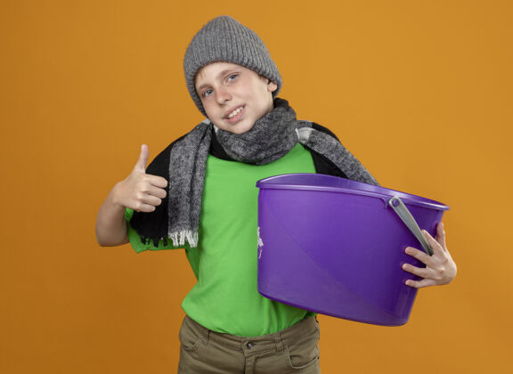 围巾生病的小男孩穿着绿色t恤 戴着暖和的围巾 戴着帽子 手里拿着垃圾 感到恶心 微笑着竖起大拇指站在橙色的墙上微笑温暖秀