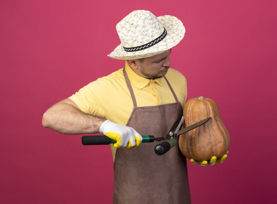 工人年轻的园丁穿着连体衣 戴着帽子 戴着工作手套 手里拿着南瓜 站在粉红色的墙上用树篱剪剪南瓜帽子年轻人篱笆