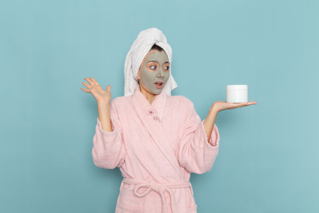 自理正面图身着粉色浴袍的年轻女性手持面霜在蓝色墙上淋浴清洗美容自理霜男性女性前面