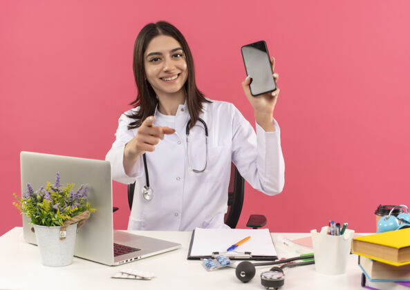 手指年轻的女医生穿着白大褂 脖子上戴着听诊器 用手指指着前面 坐在桌旁 笔记本电脑放在粉红色的墙上表演医疗保健人