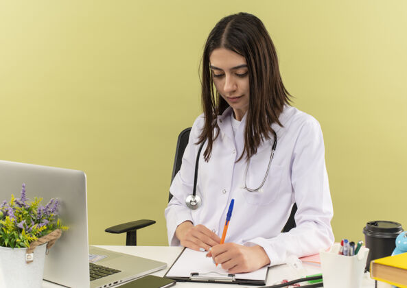 医疗保健年轻的女医生 穿着白大褂 脖子上戴着听诊器 坐在桌旁用笔记本电脑在墙上写着严肃的东西脖子人笔记本电脑