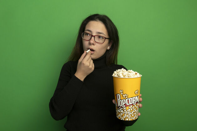 面部表情年轻漂亮的女人穿着黑色高领毛衣戴着眼镜 手里拿着一桶爆米花 一边吃着爆米花 一边开心地站在绿色的墙上抱女人黑色