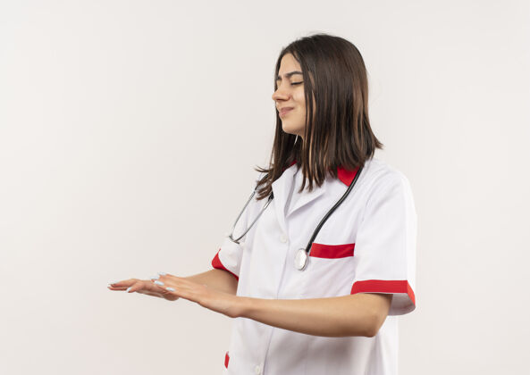 做年轻的女医生穿着白大褂 脖子上戴着听诊器 双手站在白墙上做着冷静的手势人年轻人手