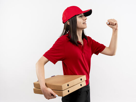 年轻年轻的送货员身穿红色制服 头戴鸭舌帽 手里拿着披萨盒 握紧拳头 站在白色的墙上 看起来很自信站着披萨送货