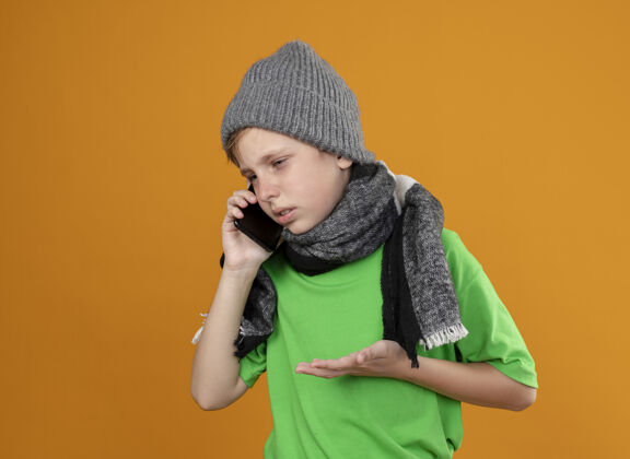 帽子生病的小男孩穿着绿色t恤 戴着暖和的围巾 戴着帽子 感觉不舒服 站在橘色的墙上讲手机 不高兴橙色手机不高兴