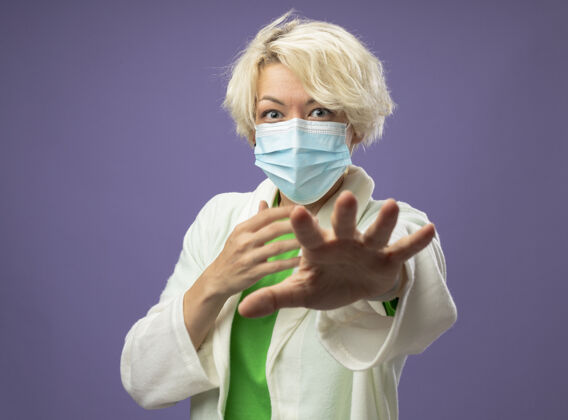 短裤生病的不健康的女人 戴着护面面具 手举紫色墙壁 做着停车标志不健康看面具