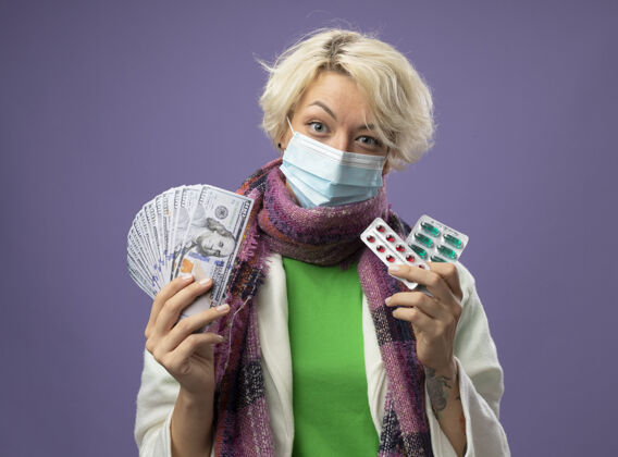 丸生病的不健康的女人 短发 戴着暖和的围巾和面罩 手里拿着现金和药水 微笑着站在紫色的墙上生病围巾保护