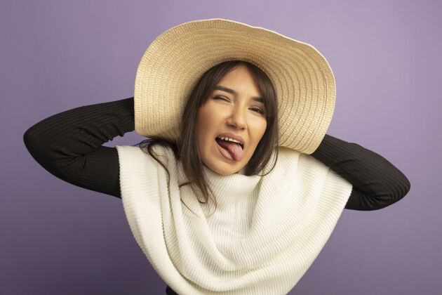 围巾戴着白领巾 戴着夏帽的年轻女子站在紫色的墙壁上 看着前面欢快地伸出舌头舌头帽子夏天
