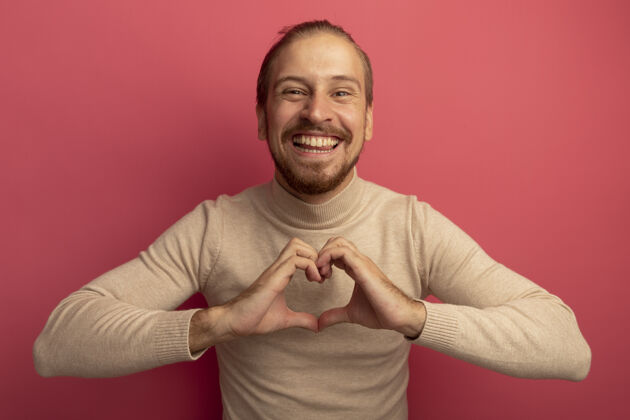 男人穿着米色套头衫的年轻帅哥站在粉色的墙上 做着胸前的心形 开心而愉快的微笑胡须人微笑
