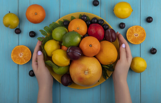 酸橙俯视图：拿着一个盘子的女人 盘子里放着混合水果 橘子 樱桃 李子 葡萄柚 柠檬 酸橙和李子 背景是绿松石色的李子保持顶部