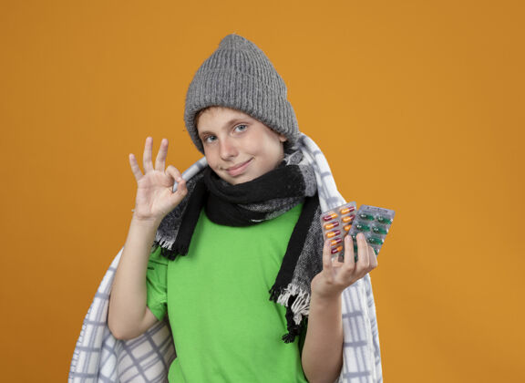 药片生病的小男孩戴着暖和的帽子 围着围巾裹在毯子里 展示着药片 感觉很好看 微笑着 展示着ok 站在橙色的墙上唱歌帽子微笑围巾