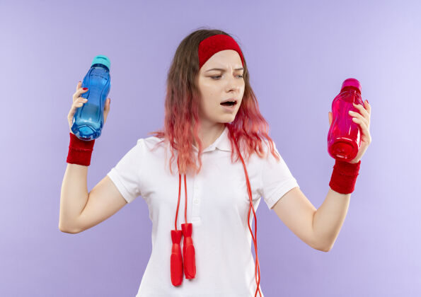 头带戴着头巾 脖子上系着跳绳 手里拿着两瓶水的年轻运动女郎站在紫色的墙上 看上去很困惑 试图做出选择人运动运动装