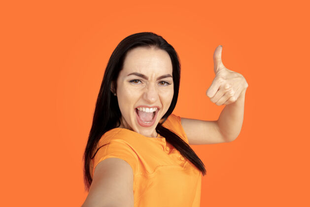 面部橙色工作室背景上的白种人年轻女子肖像穿着衬衫的漂亮黑发女性模特人类情感的概念 面部表情 销售 广告广告空间指向 展示 微笑女商人工作专业