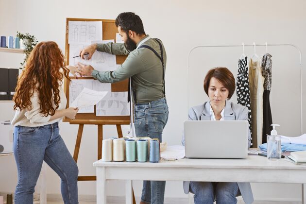 服装女时装设计师与笔记本电脑和同事在工作室工作的正面图商业裁缝线