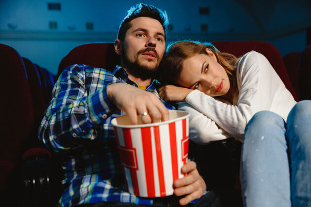 年轻迷人的年轻白种人夫妇在电影院 房子或电影院看电影看起来富于表现力 惊讶和情绪化独自坐着玩关系 爱情 家庭 周末时间电影院女室内
