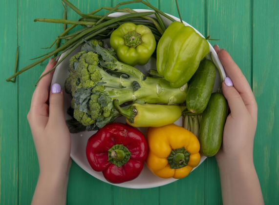 配料俯视图：一个拿着西兰花 黄瓜 彩色甜椒和葱的女人在一个绿色背景的盘子里蔬菜花椰菜洋葱