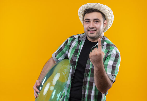 男人穿着格子衬衫 戴着夏帽 手持充气戒指 微笑着用食指站在橙色墙壁上的年轻旅行者充气旅行者姿势