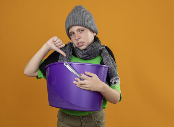 不开心生病的小男孩穿着绿色t恤 戴着暖和的围巾 戴着帽子 手里拿着垃圾 感到恶心 竖起大拇指 站在橙色的墙上不开心 生病看按住站