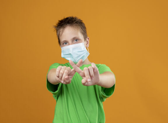 目录生病的小男孩穿着绿色t恤 戴着护面面具 做着停止手势 交叉食指站在橙色的墙上面具手势穿