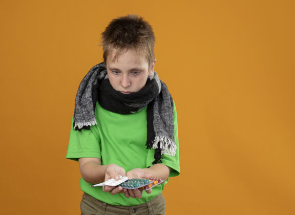 看着生病的小男孩穿着绿色t恤 脖子上围着暖和的围巾 手里拿着药片 站在橙色的墙上 看着他们被迷惑疾病男孩抱着