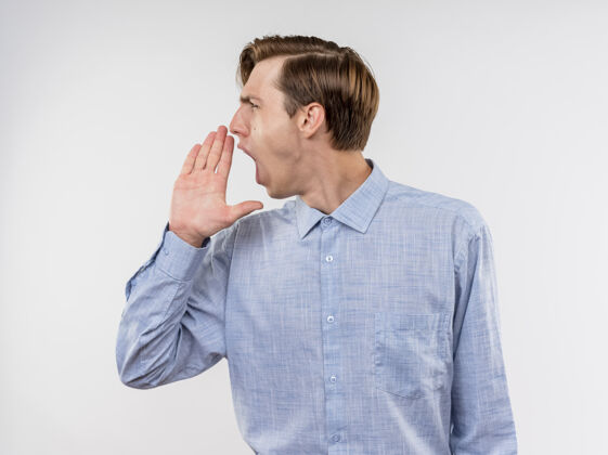 男人穿蓝色衬衫的年轻人站在白色的墙上 一边看一边用手捂着嘴大声喊叫靠边站靠近