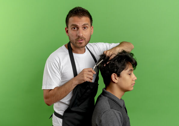 人穿着围裙的理发师用剪刀剪头发 满意的顾客站在绿色的墙上姿势站帅哥