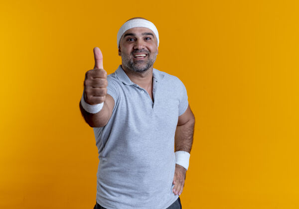 人戴着头巾的成熟运动型男人站在橙色的墙上 面带微笑 竖起大拇指站在前面脸拇指表情