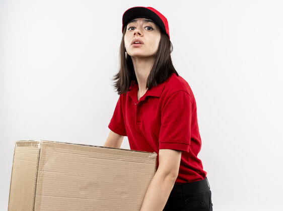 穿年轻的送货女孩穿着红色制服 戴着帽子 手里拿着一个又大又重的纸板箱 站在白色的墙上 看上去很累重送货累了