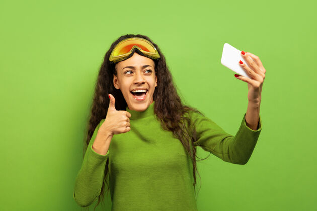 女性以绿色工作室为背景 黑色皮肤的非洲裔美国年轻女子滑雪面具肖像关于人类情感 面部表情 销售 广告 冬季运动和假期的概念自拍或虚拟博客美国微笑欢呼