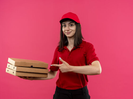 盒子身穿红色马球衫 头戴鸭舌帽的年轻送货女孩拿着披萨盒 用食指指着他们友好地微笑着站在粉色的墙上年轻拿着帽子