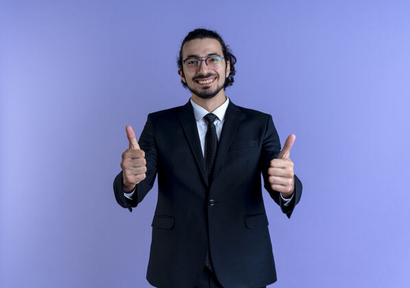 男人一个穿着黑色西装戴着眼镜的商人微笑着望着前面 竖起大拇指站在蓝色的墙上看站拇指