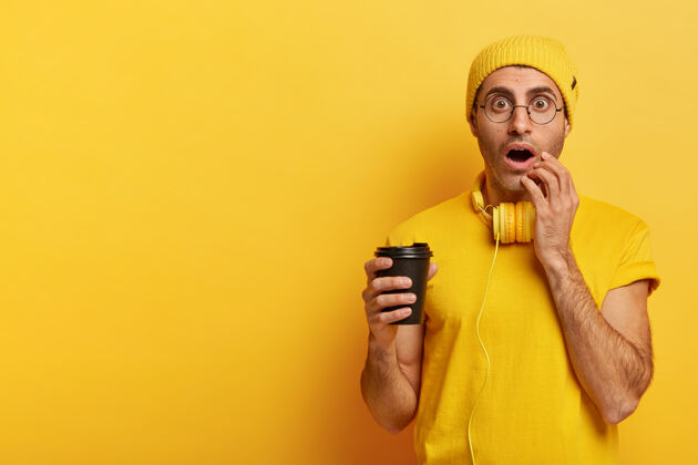 男人情绪化的年轻人拿着一次性咖啡 睁大眼睛盯着 不敢相信的眼睛 戴着黄色的帽子饮料千禧一代惊呆
