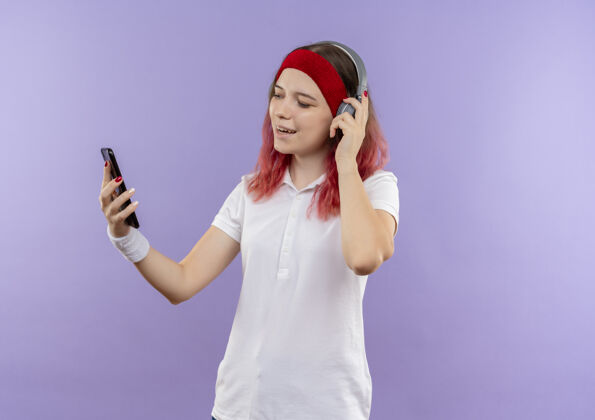运动装站在紫色的墙上 戴着耳机 看着智能手机屏幕上微笑着听音乐的年轻运动女性年轻人看手机