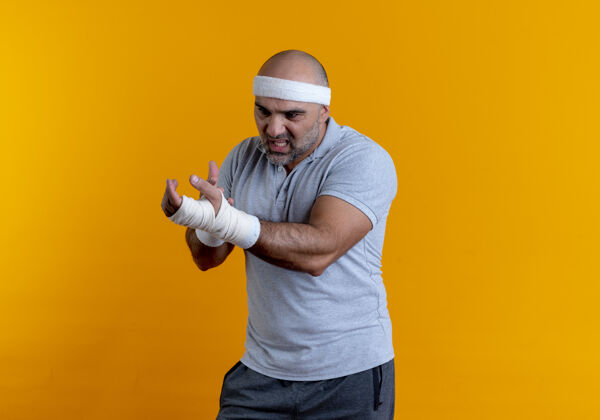 绷带戴着头巾的成熟的运动型男人站在橘色的墙上 摸着他缠着绷带的手 看起来很不舒服手运动成熟