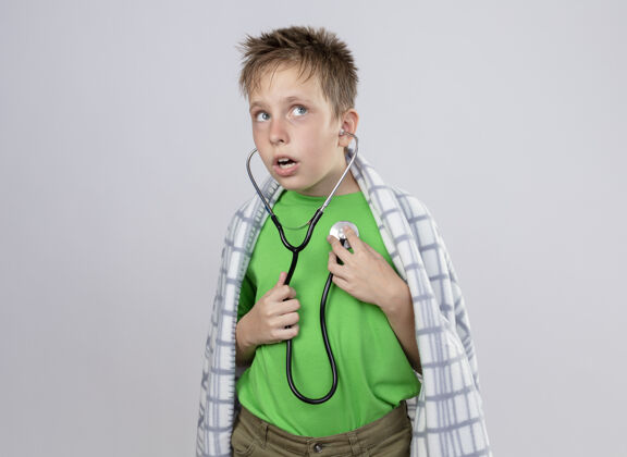 不适生病的小男孩穿着绿色t恤 裹着毛毯 脖子上戴着听诊器 站在白色的墙上 听着心跳混乱小毯子男孩