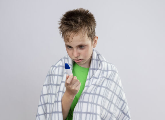 小生病的小男孩穿着绿色t恤 裹着毛毯 看着温度计 不安地站在白色的墙上看病担心