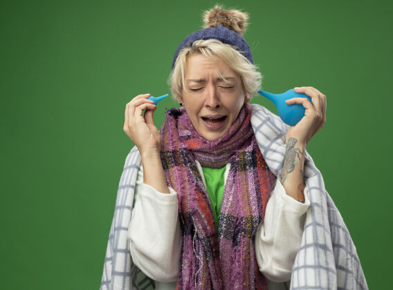 毯子生病的不健康的女人 短头发 戴着暖和的围巾 戴着帽子 感觉不舒服 裹着毯子 拿着灌肠剂 脸上带着恼怒的表情 站在绿色的墙边不适头发烦躁