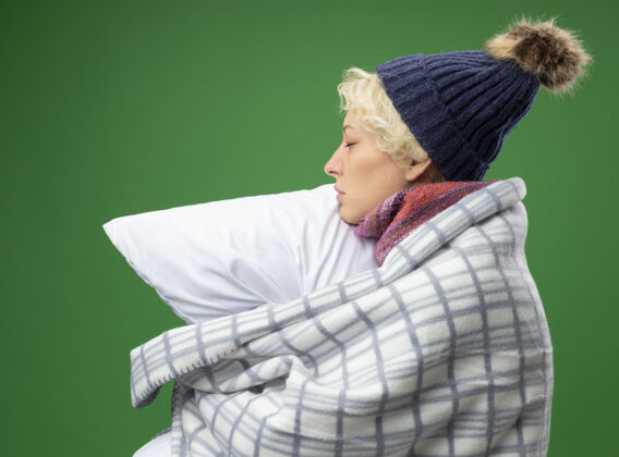 不适生病的不健康的女人 短头发 戴着暖和的围巾和帽子 感觉不舒服 裹着毯子 抱着枕头 侧着身子站在紫色的墙上不健康短裤女人