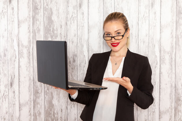 正面正面图穿着严苛衣服的年轻女商人黑色夹克在白色墙壁上用她的笔记本电脑带着微微的微笑工作办公室的女商人女风景西装