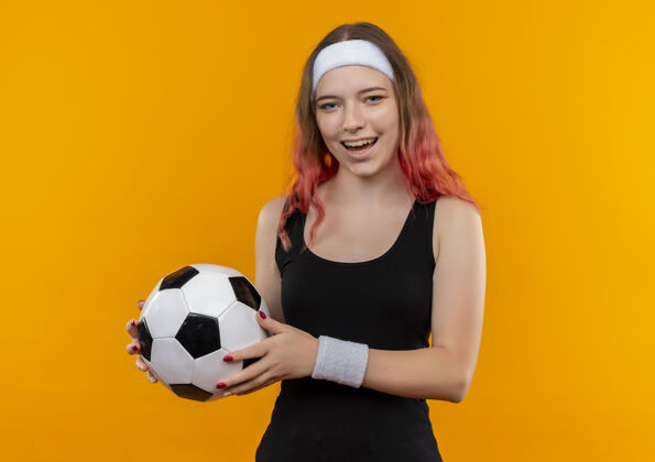 抱身着运动服的年轻健身女士 手拿足球 脸上洋溢着幸福的笑容 站在橙色的墙上看人快乐