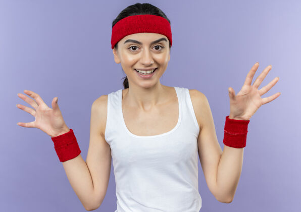 年轻身穿运动服的年轻健身女 头戴束带 双手举着投降 微笑着站在紫色的墙上市民健康举起