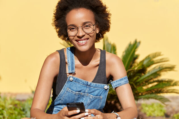 牙齿笑容可掬 戴圆眼镜 使用智能手机进行在线通讯 连接无线互联网 在热带地区休息的满意黑人女性的后视图科技理念电话小工具眼镜