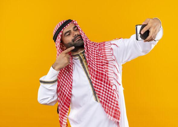 站身着传统服装的阿拉伯商人站在橙色的墙上 用智能手机自拍 看上去很自信姿势人公民