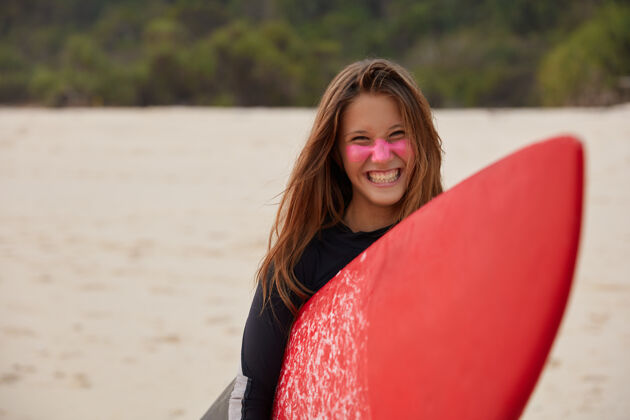 站立开朗的职业年轻女冲浪者笑容灿烂 扛着红蜡冲浪板 脸上有锌 打了大海浪后高兴 靠着沙墙造型 表现积极游泳海滩姿势