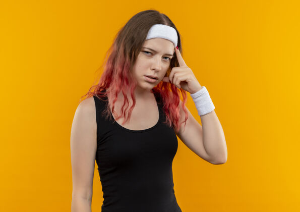 站身着运动服的年轻健身女士指着太阳穴 站在橘色的墙上 看起来很自信集中运动装人
